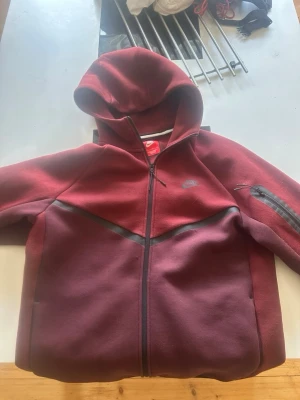 Bordeaux Nike hoodie med dragkedja - Snygg hoodie från Nike i bordeauxrött med svarta detaljer och hel dragkedja framtill. Hoodien har en huva och en praktisk ficka med dragkedja på ärmen. Perfekt för en sportig och avslappnad stil. Hämtas upp vid dörren eller nära min stad 