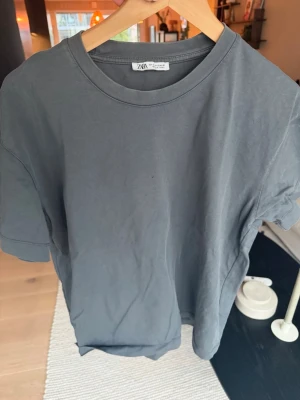 Mörkgrå basic t-shirt från ZARA - En enkel och stilren mörkgrå t-shirt från ZARA i mjuk bomull. Klassisk passform med rund halsringning och korta ärmar. Perfekt till jeans eller shorts för en clean look.