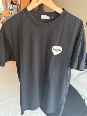 Svart Carhartt WIP t-shirt med hjärta - Snygg svart t-shirt från Carhartt WIP med ett vitt hjärta och logga tryckt på bröstet. Klassisk rund halsringning och korta ärmar. Tillverkad i mjuk bomull som känns skön mot huden. Perfekt för en avslappnad streetwear-look.
