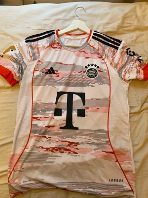 Bayern München Müller matchtröja - Säljer en vit och röd Bayern München matchtröja från Adidas med svart logga och stora T-trycket på bröstet. På ryggen står det Müller 25 med grafik av spelaren och FC Bayern München-tryck. Tröjan har korta ärmar och AEROREADY-material för bra ventilation.