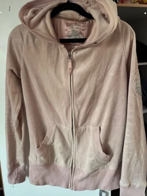 Ljusrosa hoodie från Greenwich Village - Ljusrosa hoodie från Greenwich Village med dragkedja framtill, två fickor och ribbade muddar. Tillverkad i mjuk bomull och har en broderad detalj på ärmen. Perfekt för en avslappnad och chill stil.