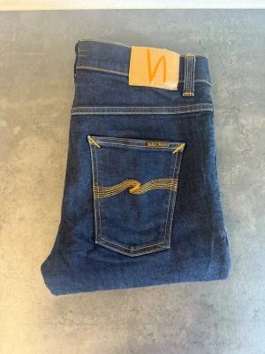 Nudie Jeans Lean Dean, stl W32 L32 - Säljer ett par mörkblå jeans från Nudie Jeans med klassisk femficksmodell och gul kontrastsöm. Jeansen har rak passform och är tillverkade i kraftigt denimtyg. Snygg detalj med Nudie-loggan på bakfickan och läderpatch i midjan.