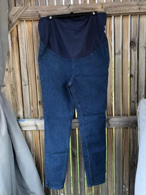 Mammabyxor i mörkblå denim - Mörkblå mammabyxor i stretchig denim med hög, mjuk mudd över magen för extra komfort. Byxorna har smal passform och klassisk jeanslook utan synliga fickor framtill. Perfekta för dig som vill ha både stil och bekvämlighet under graviditeten.