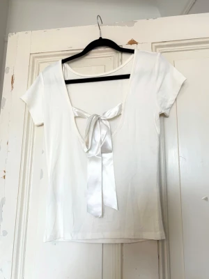 Vit topp med öppen rygg och rosett - En jättefin vit t-shirt som har öppen rygg med en satinrosett baktill. Storlek S. Helt ny med prislappen kvar! Kontakta vid frågor💕
