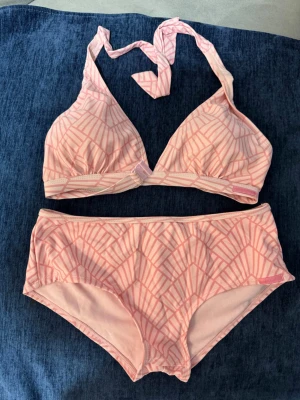 Rosa bikini med grafiskt mönster - Säljer en söt bikini i ljusrosa med grafiskt mönster i en mörkare rosa nyans. Överdelen har halterneck-knäppning och vadderade kupor, medan underdelen har en högre midja för extra komfort. Perfekt för strandhäng eller poolen!