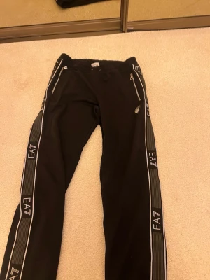 Svarta mjukisbyxor från EA7 Armani - Säljer ett par svarta mjukisbyxor från EA7 Emporio Armani med vita detaljer och dragkedjor vid fickorna. Byxorna har breda revärer längs benen med EA7-logga och resår i midjan. Perfekta för en avslappnad och sportig stil.