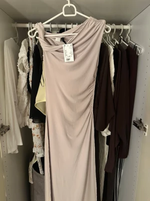 Ljusbeige långklänning med drapering - Supersnygg långklänning i ljusbeige med draperad halsringning och lätt rynkade detaljer längs sidan. Klänningen har en elegant, figurnära passform och är ärmlös. Perfekt för dig som vill sticka ut med en stilren look.