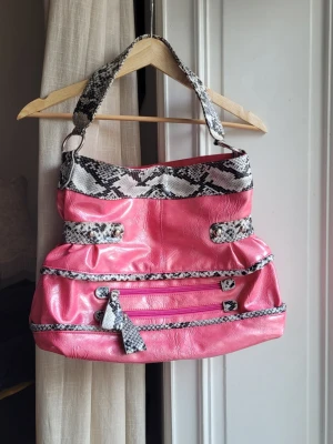 Rosa handväska med ormmönstrade detaljer y2k - Cool rosa handväska i glansigt material med ormmönstrade detaljer på handtag, kanter och dragkedjor. Väskan har flera fack framtill och en bred axelrem. Perfekt för dig som vill sticka ut med en färgglad accessoar. Svart fläck under väskan i övrigt bra skick 