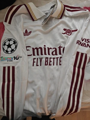 Vit Arsenal matchtröja Adidas spelar Version  - Säljer en vit Arsenal matchtröja från Adidas med vinröda detaljer och ränder på ärmarna. Tröjan har klubbmärke, Adidas-logga och sponsortryck framtill samt Champions League-märke på ärmen. Stort nummer 29 i vinrött med guldkant på ryggen. Klassisk pikékrage.