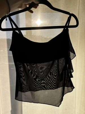 Svart transparent volangtop - Säljer en svart, transparent festtopp med tunna justerbara axelband och volangdetaljer framtill och på sidan. Toppen är luftig och har en flowy passform, perfekt för att styla lager på lager eller till ett par jeans eller kjol! Extra tyg framför bysten som ej är genomskinligt. Har tyvärr aldrig använt, även om den är skitcool, har bara för mycket grejer:/