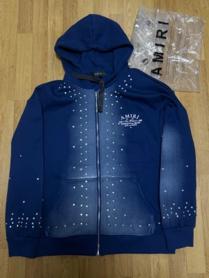 Amiri Zip Hoodie with stones – Size M - Zip Hoodien är helt ny, inga skador eller slitage. Originalförpackning medföljer. Exklusiv zip hoodie från Amiri med dekorativa stenar på fram- och baksida samt ärmar. Stor logotyp på ryggen, tryck på bröstet och en stilren design som sticker ut.  Size: M 🏷️ Rabatt vid köp av flera plagg!