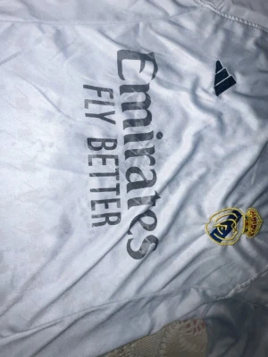 Real Madrid vit matchtröja Adidas - Vit Real Madrid matchtröja från Adidas med klubbmärke på bröstet och 'Emirates Fly Better' tryck på framsidan. Tröjan har svarta ränder på axlarna och är gjord i ett lätt, sportigt material som andas. Perfekt för fotboll eller träning.
