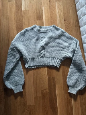 Grå cropped stickad tröja - Trendig grå cropped stickad tröja med breda ärmar och ribbade muddar. Tröjan har ett snyggt tvinnat kabelmönster framtill och är gjord i ett mjukt material som känns skönt mot huden. Perfekt för lager på lager och en cool vardagslook.