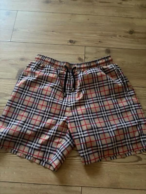 Rutig shorts från Burberry - Snygga shorts från Burberry med klassiskt rutigt mönster i beige, svart, vitt och rött. De har elastisk midja med snörning och är perfekta för sommaren. Lätta och bekväma med meshfoder på insidan.
