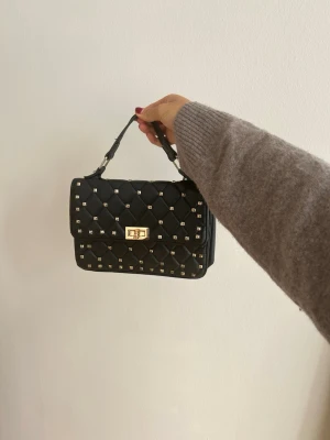 Valentino likande väska med nitar💗💗💗 - Snygg svart handväska i quiltat mönster med små silvriga nitar och guldfärgat vridlås. Väskan har ett kort handtag och en boxig form, perfekt för att lyfta din outfit. Materialet ser ut som konstläder.