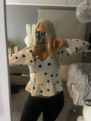 Vit blus med svarta prickar och v-ringning - Säljer en vit blus med stora svarta prickar och v-ringad hals. Blusen har lång ärm och en markerad midja med volang nedtill som ger en snygg siluett. Perfekt för dig som gillar trendiga mönster och en feminin look.