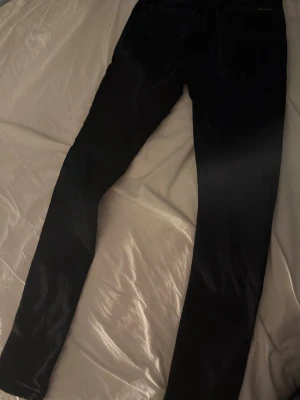 Svarta jeans från Nudie - Säljer ett par mörkblå skinny jeans från Nudie Jeans med klassisk femficksmodell och läderpatch bak i midjan. Jeansen har smal passform och är tillverkade i stretchigt denimtyg för extra komfort. Perfekta för en clean och stilren look.