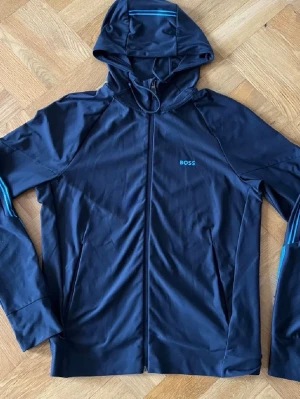 Mörkblå boss hoodie  - Snygg mörkblå hoodie från BOSS med dragkedja framtill och huva. Diskret BOSS-logga i blått på bröstet och tunna blå ränder på ärmarna. Tillverkad i ett mjukt och stretchigt material som känns skönt mot huden. Perfekt för en avslappnad och sportig stil.