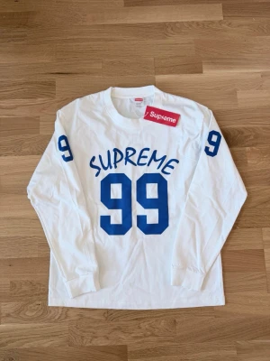 Vit långärmad Supreme topp - Vit långärmad topp från Supreme med blå tryckta siffror '99' på bröstet och ärmarna samt Supreme-logga. Oversized fit och rund halsringning. Perfekt för dig som gillar streetwear och vill sticka ut med en sportig vibe.