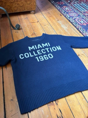 Mörkblå stickad tröja MIAMI 1960 - Mörkblå stickad tröja med texten 'MIAMI COLLECTION 1960' i vitt framtill. Tröjan har rund halsringning, långa ärmar och breda ribbade muddar vid ärmslut och nederkant. Perfekt för dig som gillar retrostil och statementplagg. Passar S/XS.