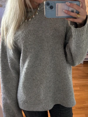 Grå stickad hoodie - Mysig grå stickad hoodie med avslappnad passform och långa ärmar. Tröjan har en huva och är tillverkad i ett mjukt, tjockt material som passar perfekt för kyliga dagar. Enkel och stilren design utan synliga loggor eller mönster.