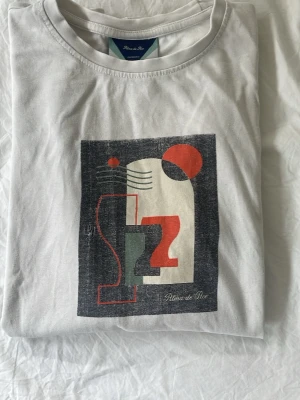 Vit t-shirt med grafiskt tryck - Vit t-shirt från Plans de Rue med ett stort grafiskt tryck i svart, rött och grönt på bröstet. Klassisk rund hals och korta ärmar. Perfekt för dig som gillar modern och stilren design.