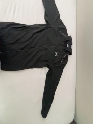 Svart windbreaker från Under Armour  - Säljer en svart hoodie från Under Armour med lång ärm och huva. Framtill finns en liten logga och på ryggen står det 'UNDER ARMOUR' i vitt. Tillverkad i ett lätt och sportigt material, perfekt för träning eller chill. Den passar även L ifall man har den storleken😁