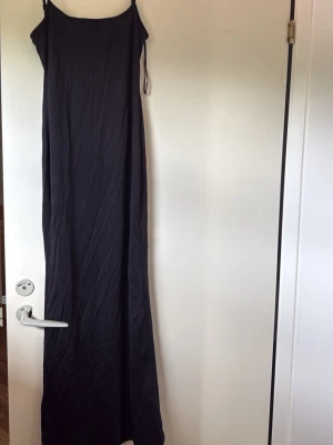 Svart långklänning från Bershka - En stilren svart långklänning från Bershka med tunna axelband och rak passform. Klänningen är enkel och minimalistisk, perfekt för dig som gillar en clean look. Tillverkad i ett mjukt och följsamt material som känns skönt mot huden.