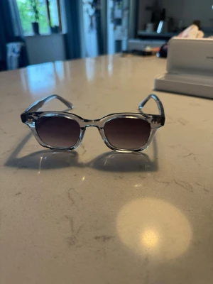 Chimi 02  - Snygga solglasögon med transparent båge och runda, mörktonade glas. Perfekt accessoar för soliga dagar och ger en cool vibe till din outfit. Lätta och bekväma att bära hela dagen.