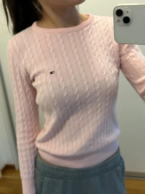 Ljusrosa kabelstickad tröja Tommy Hilfiger - Säljer en ljusrosa kabelstickad tröja från Tommy Hilfiger med rund halsringning och diskret logga på bröstet. Tröjan har långa ärmar och ribbade muddar vid ärmslut och nederkant. Perfekt för dig som gillar klassisk och stilren look.
