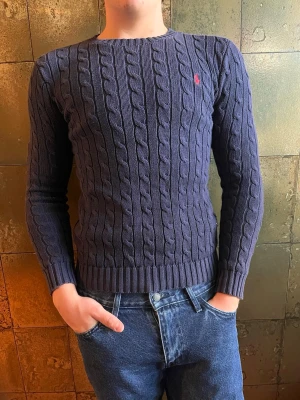 Mörkblå kabelstickad tröja Polo Ralph Lauren - Säljer en mörkblå kabelstickad tröja från Polo Ralph Lauren med klassisk röd logga broderad på bröstet. Tröjan har rund halsringning, långa ärmar och ribbade muddar. Perfekt för dig som gillar stilrena och tidlösa plagg.
