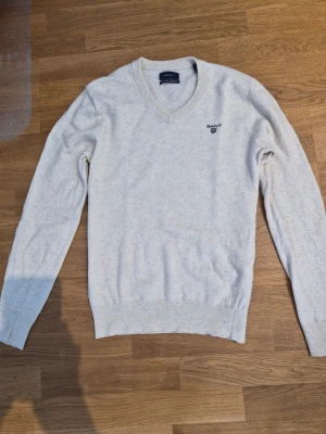 Beige stickad GANT-tröja - Säljer en beige stickad tröja från GANT med V-ringning och broderad logga på bröstet. Eftersom den krympt i tvätten en gång passar den nog bättre som storlek S!