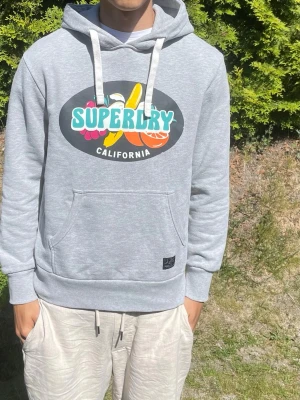 Grå Superdry hoodie - Snygg grå hoodie från Superdry med färgglatt tryck på bröstet där det står 'Superdry California' och fruktmotiv. Hoodien har vit dragsko i huvan och en stor magficka. Tillverkad i mjukt bomullsmaterialSuperdry Hoodie  Säljer min Superdry hoodie i fint skick. Bekväm passform och perfekt för både vardag och fritid.  📦 Snabb frakt ✅ Bra skick 💯 Äkta Superdry  Storlek: S  #superdry #hoodie #superdryhoodie #streetwear #fashion #mode #vinted #plick #sweater #hooded #casual #trend #Grisch