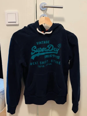 Mörkblå Superdry hoodie med tryck - Snygg mörkblå hoodie från Superdry med turkost tryck på bröstet och text på både engelska och japanska. Klassisk känguruficka framtill, ribbade muddar och justerbar huva med vita snören. Perfekt för en avslappnad streetwear-look. Skriv till om ni är intresserade 😁