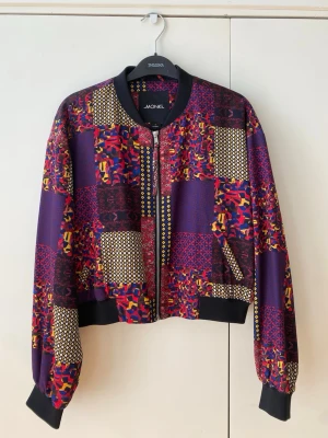 Mönstrad bomberjacka från Monki - Färgstark bomberjacka från Monki med patchwork-mönster i lila, rött, gult och svart. Jackan har svarta muddar vid ärmslut och midja, dragkedja framtill och rundad krage. Materialet är mjukt och lätt, perfekt för att lyfta din outfit med en unik stil.