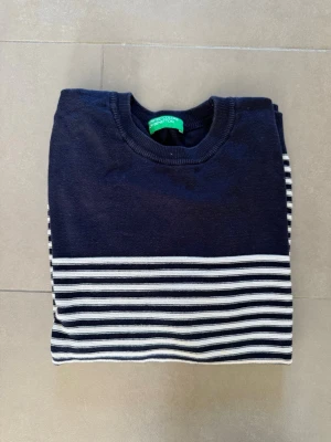 Marinblå randig tröja Benetton - Snygg marinblå stickad tröja från Benetton med vita horisontella ränder på nedre delen och ärmarna. Tröjan har rund halsringning och ribbade muddar vid ärmslut och nederkant. Perfekt för en stilren och avslappnad look.