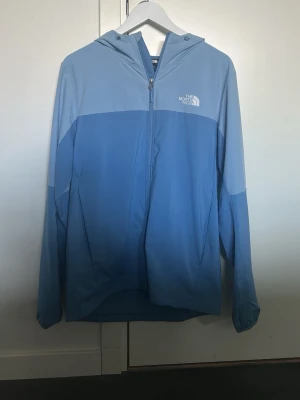 Blå vindjacka från The North Face - Snygg tvåtonad blå Windbreaker från The North Face med huva och hel dragkedja framtill. Jackan har reflekterande detaljer på ärmen och bröstet samt elastiska muddar vid ärmsluten. Perfekt för blåsiga dagar och utomhusaktiviteter.
