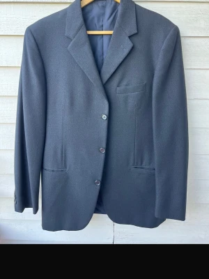 Svartblå - Stilren svartblå kavaj med tre knappar framtill och klassiska slag. Kavajen har två framfickor och en bröstficka. Materialet ser ut att vara en slät ullblandning som ger en elegant look. Perfekt för dig som vill ha en enkel och tidlös stil.