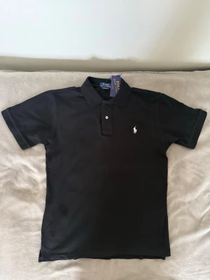 Svart pikétröja från Ralph Lauren - Klassisk svart pikétröja från Ralph Lauren med vit broderad logga på bröstet. Tröjan har korta ärmar, krage och knappar framtill. Tillverkad i bomull för en bekväm och stilren look.