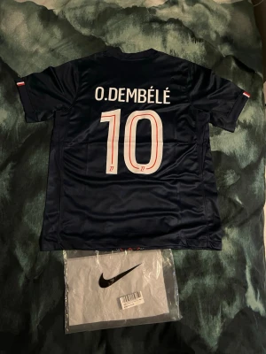 PSG matchtröja O. Dembélé #10 Nike - Snygg mörkblå PSG matchtröja från Nike med O. Dembélé och nummer 10 på ryggen. Framsidan har Qatar Airways-logga och PSG-emblem, samt röd och vit detalj längs mitten. Tillverkad i lätt och ventilerande material, perfekt för fotboll eller träning.