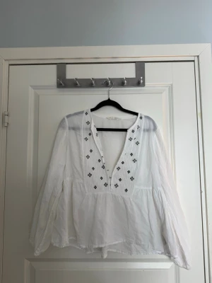 Vit odd molly blus med broderi - Säljer en vit odd molly blus med v-ringning och svarta broderade detaljer framtill. Blusen har  lång ärm och är tillverkad i ett tunt, luftigt material som ger en avslappnad och flowy känsla. Perfekt för dig som gillar en chill och trendig stil. Storlek S