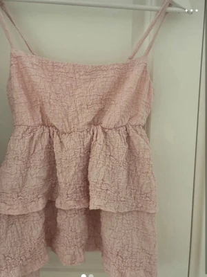 Ljusrosa volangtop från Gina Tricot - Supersöt ljusrosa topp från Gina Tricot med tunna axelband och volangdetaljer. Toppen har en skrynklig struktur och är perfekt för varma dagar. Passar dig som gillar en feminin och luftig stil.