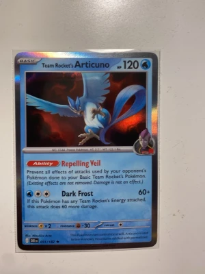 Pokémonkort Team Rocket's Articuno - Säljer ett Team Rocket's Articuno Pokémonkort med holografisk yta. Kortet har blå och silver färg, med Articuno i fokus och Team Rocket-tema. Kortet är gjort av laminerad kartong och har coola attacker som Repelling Veil och Dark Frost.