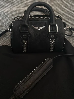Svart handväska Zadig & Voltaire - Cool svart handväska i läder från Zadig & Voltaire med silverfärgade detaljer, nitar längs kanterna och dubbla dragkedjor framtill. Väskan har ett emblem med vingar och justerbar axelrem med nitar. Perfekt för dig som gillar edgy och stilren design.