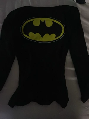 Svart Batman långärmad tröja - Cool långärmad svart tröja med stort gult Batman-logga tryck på bröstet. Perfekt för cosplay, maskerad eller för dig som älskar superhjältar. Tröjan har en klassisk passform och är tillverkad i mjukt material.