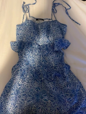 Blåvit blommig klänning Gina Tricot - Söt blåvit klänning från Gina Tricot med blommigt mönster och volangdetaljer. Klänningen har smala axelband som knyts och är tillverkad i ett lätt bomullstyg. Perfekt för varma dagar och somriga vibes!