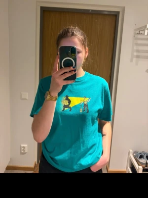 Turkos t-shirt med tryck - Säljer en turkos t-shirt med coolt grafiskt tryck framtill i gult, svart och guld. T-shirten har normal passform och korta ärmar. Perfekt för dig som vill ha en färgglad och unik look.