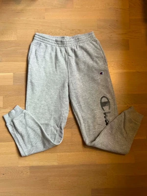 Gråa mjukisbyxor från Champion - Snygga gråa mjukisbyxor från Champion med elastisk midja och muddar vid bensluten. Byxorna har en liten Champion-logga på låret och stor logga längs benet. Perfekta för chill eller träning. Tillverkade i mjukt sweatshirtmaterial.