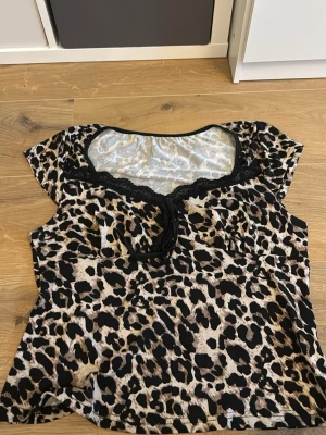 Leopardmönstrad topp med spets - Snygg leopardmönstrad topp med korta ärmar och svart spetsdetalj vid urringningen. Toppen har en liten rosett framtill och är tillverkad i ett mjukt, lätt material som känns skönt mot huden. Perfekt för dig som vill sticka ut med ett djurmönster.