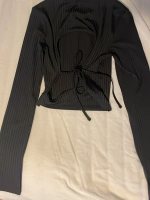Svart ribbad långärmad topp med knyt - Trendig svart ribbad topp med lång ärm och snygg knytning framtill. Croppad modell som ger en cool look, perfekt att styla med höga jeans eller kjol. Materialet är stretchigt och bekvämt.